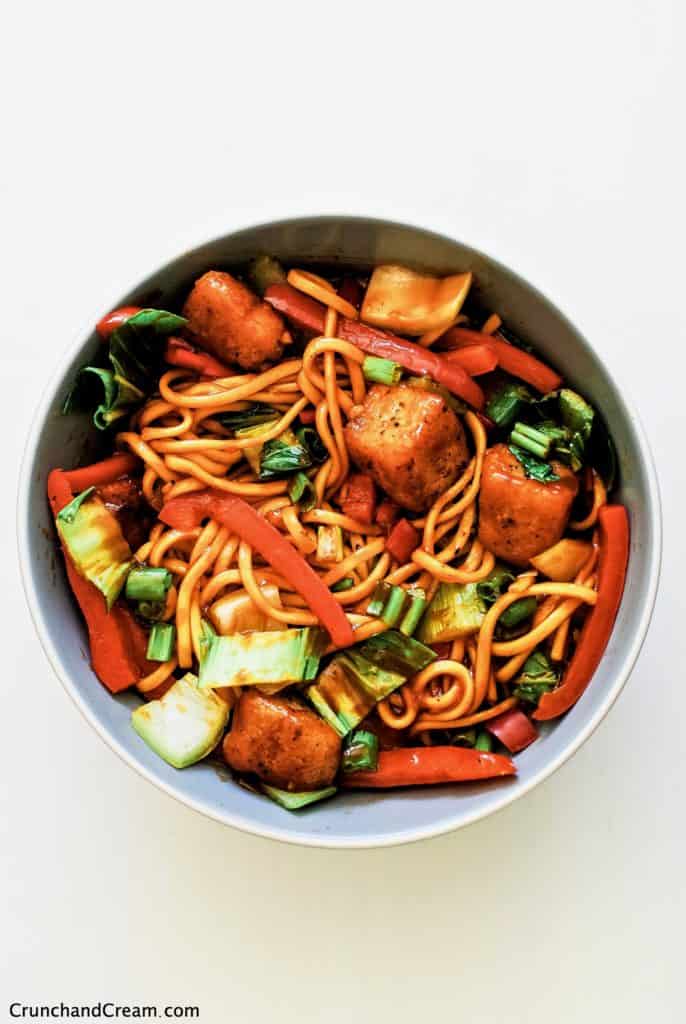 overhead of a deep bowl full of fresh egg noodles, red bell pepper, pak choi and spring onions with crispy diced halloumi pieces in a spicy sweet chilli sauce
