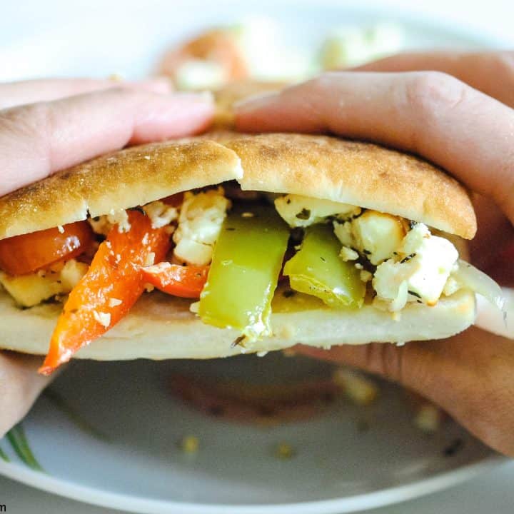 hands holding a bread roll filled with peppers, tomatoes, onions and feta