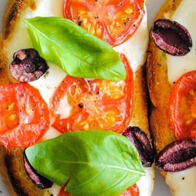 close-up of toast topped with melted mozzarella, sliced tomatoes, halved olives and basil leaves