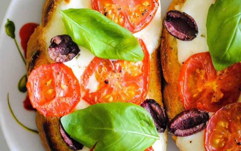 close-up of toast topped with melted mozzarella, sliced tomatoes, halved olives and basil leaves