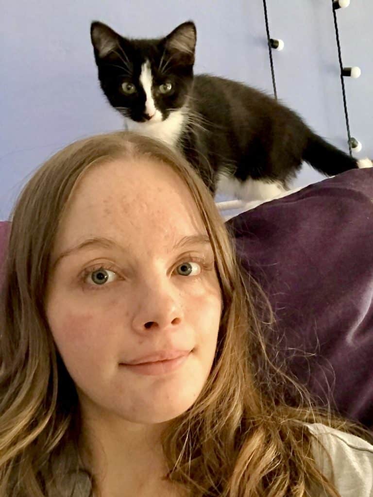 Photo of a smiling 18-year-old with brown hair and a black and white cat behind her