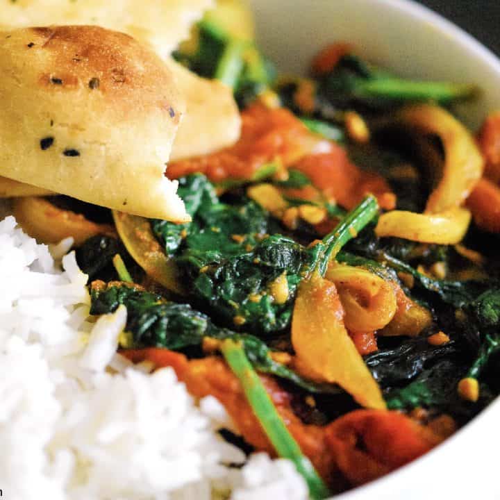 a close-up shot of a vegetable masala curry in a bowl with spinach, onions and tomatoes. Served with basmati rice and a torn naan bread.